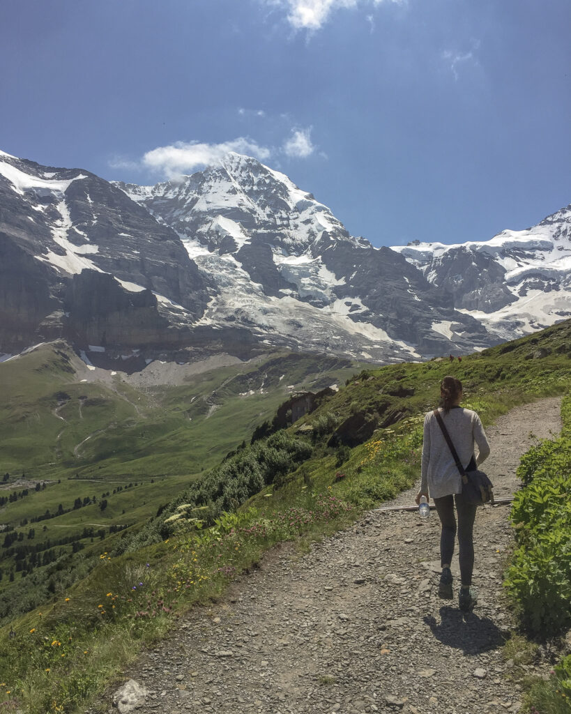 Amy hikes at the top of the Swiss Alps on The Royal Walk, Switzerland. Lush green mountains and snow-capped peaks provide a breathtaking view.